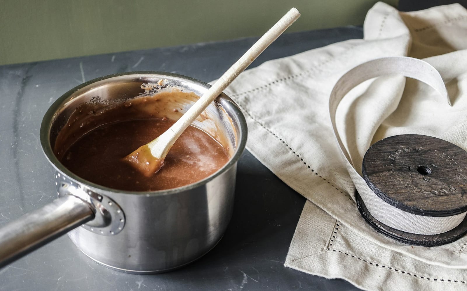 Panela de metal com chocolate derretido e uma colher de madeira ao lado de um pano de linho